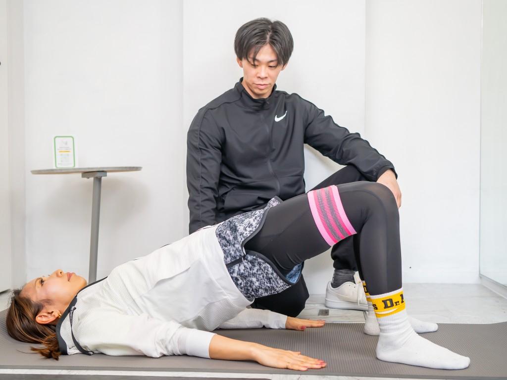 マットの上でヒップブリッジを行い、太ももにレジスタンスバンドを巻いた女性と、フォームを見守るトレーナー。